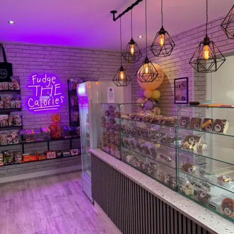 Want That Fudge UK The interior of the Want That Fudge shop in Scunthorpe. There is a neon sign on the wall and shelves featuring a selection of fudge, sweets and chocolate.