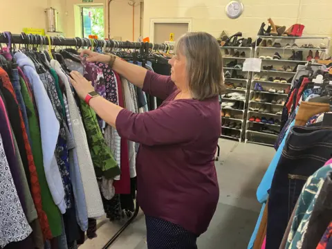 Amy Holmes/BBC A picture of a woman with a wine-red coloured top and brown hair looking through a selection of clothes on a hanger.