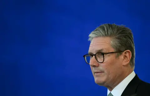 Reuters A grey-haired man in thick-rimmed dark glasses looking into the centre of the frame. The background is a deep blue wall