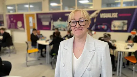 Woman with blonde hair, dark glasses, grey blazer, white tee-shirt and silver necklace.

Standing up for the photo with the classroom in the background.