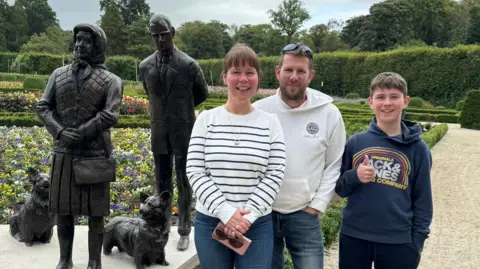 The family of three standing in front of the sculptures. 

Cheryl is wearing a white stripy jumper, blue jeans and she is carrying a pink purse and brown pair of sunglasses. She has brown hair and a  fringe. She is smiling. 

Gordon is wearing sunglasses on his head. He has brown hair and a subtle beard. He is wearing a white hoody and blue jeans. He is smiling. 

Their son is wearing a navy Jack & Jones hoody and black trousers. He has short brown hair and is putting his thumb up, smiling. 