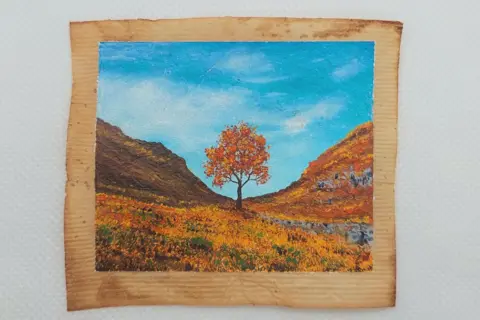 Caroline West The Sycamore Gap tree on a used teabag painting in an Autumn setting. It has orange leaves and the field around it is in Autumnal colours.