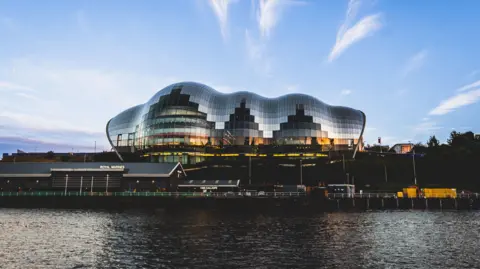 Tyneside Photographic An image of the glasshouse international centre for music which has reflective panels on the outside. In the foreground are some smaller buildings and a river. In the background there's a blue sky with some light clouds. The photo has been taken at dusk so the light is fading.