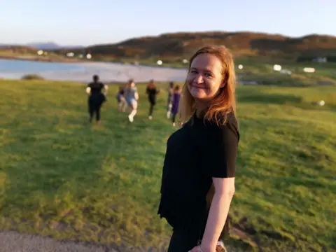 Christopher MacRae Dr MacRae turning to look at the camera, wearing black clothing. She is standing in front of an area of grass, with Mellon Udrigle beach in the distance. There is a group of young people walking on the grass.