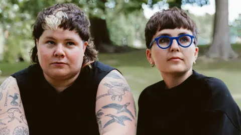 Rosie Powell ARXX, made up of (L-R) Hanni Pidduck and Clara Townsend. Hanni has short brown hair with a blonde streak in their fringe and has heavily tattooed arms. Clara also has short brown hair with a fringe and wears cobalt blue rimmed round glasses. They're both dressed in black and are pictured sitting outside in a woodland area.
