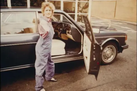 Chalk Productions/Alison Lapper Alison Lapper, wearing a red t-shirt and blue dungarees, stands in front of a black car. The driver's seat of the car is open. 