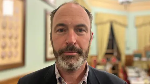 Andrew Cross in the council chamber. He has a beard and is wearing a suit jacket.