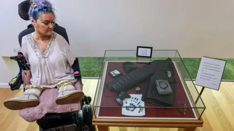 Alamy Alison Lapper looks at a collection of her son's belongings on a table next to her. The belongings include his coat, bag and games console. Alison is wearing a pink top and has purple dyed hair.