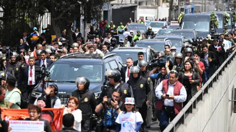 Getty Images The hearse carrying the body of former president Alberto Fujimori is flanked by security, police, reporters and camera crews as a convoy of black vehicles make their way towards his funeral service,