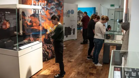 Martin Rowe/Peterborough Museum and Art Gallery A woman in a dark top, skirt and tights, looking at the contents of an exhibition case, in front of a large display showing an artist's impression of a Bronze Age settlement going up in flames, with two men and two women looking at other displays at Peterborough Museum