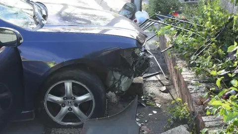 Avon and Somerset Police The front of the car which has been mounted onto the pavement. The bumper is smashed and crumpled from the force of the impact. There is also an old bed frame which has been crumpled and is lying next to the low brick wall which Mr Tjitendero was pinned against.