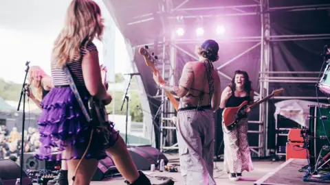 Derek Bremner Panic Shack performing on stage at a festival. Megan, pictured on the right, smiles widely at her bandmates while holding her guitar. Bassist Emily Smith and guitarist Romi Lawrence are pictured with their backs to the camera while singer Sarah Harvey is at the top of the stage singing to the crowd. 