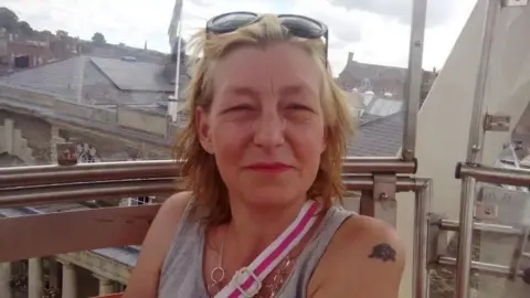 Facebook Dawn Sturgess has short blonde hair and dark sunglasses on her head, She is wearing a grey vest top, silver chain necklace and the pink and white strap of bag is crossing her body. She is sitting on a balcony with rooftops visible in the background