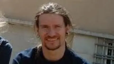 Kate Wilson Mark Kennedy wearing a t-shirt and smiling with a building behind him