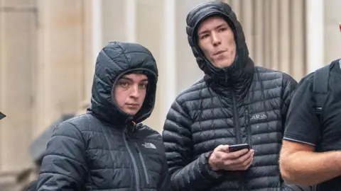 PA Media Two men in black coats, stood with their hoods up outside a court building. One of them is holding a mobile phone.