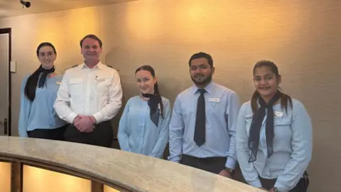 Five members of staff standing side by side. Four are dressed in a blue shirt with a black tie or black scarf. The captain is between them dressed in a white shirt with black trousers.