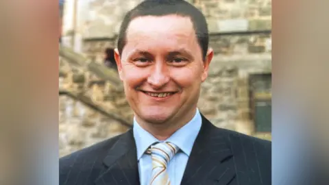 John Kelly  A younger John Kelly wearing a suit with a blue shirt and multi-coloured stripey tie. He has dark hair and is smiling at the camera