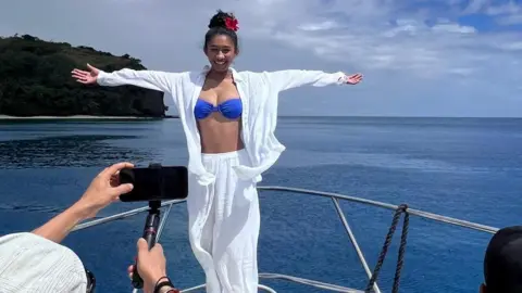 JENNIFER CHAN A woman wearing a white shirt and bottoms and blue bikini top poses on a boat in the sea, in front of a man picturing a picture with his phone.