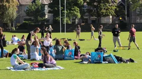 Aberdeen University Students' Association Groups of young people sitting on the grass in the sunshine - some lying down, some with delivery bags of Dominos pizza, others playing a ball game in the background