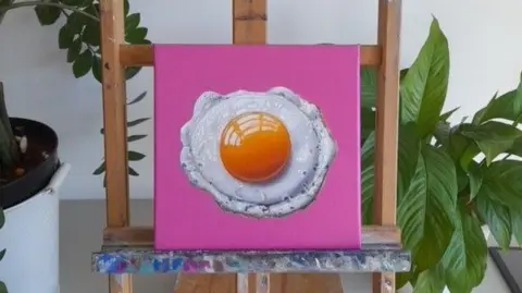 Daniel Halksworth A sunny-side up on a pink canvas, popped on a small easel with paint marks on the wood. Two plants can be seen in the background.