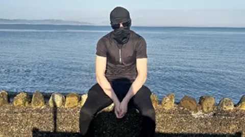  Counter Terrorism Policing Wales Alex Hutton, who has been jailed for more than five years, sits on a wall in front of the sea while wearing a balaclava and sunglasses