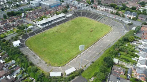 PA Media An aerial view of a grass pitch with concrete terrace-style stands. It sits in a residential area with a number of red brick houses surrounding the site. 