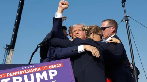 Getty Images Donald Trump, surrounded by Secret Service agents wearing dark glasses who hold him close to them, raising a fist in defiance with a streak of blood across his face