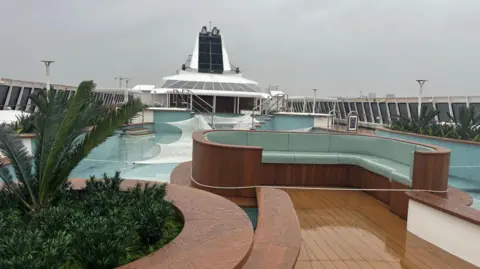 A wooden seating area with blue cushions is on the deck. Behind this is a pool with steps into it.