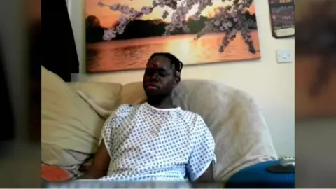 Avon and Somerset Police Katungua Tjitendero sat on a sofa wearing a hospital gown. There is a large painting of a lake on the wall behind him. 