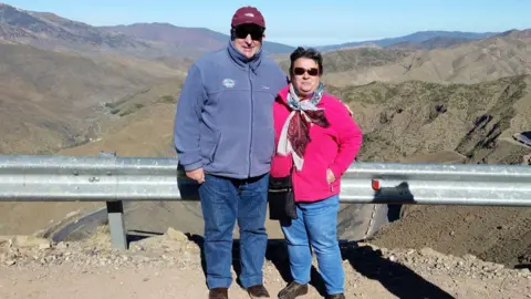 Contributed Rodrick Lodge and his wife Pauline standing in front of a mountainous landscape. They are both wearing fleeces and sunglasses and smiling at the camera. 