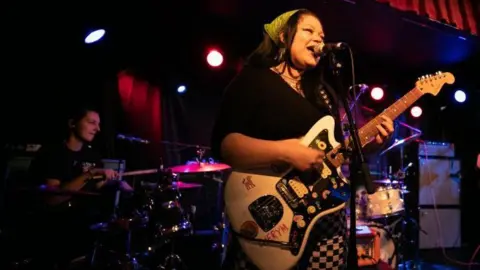 Getty Images Cherym performing on stage in Dublin in 2023. Hannah sings into a microphone at the front of the stage, holding a white electric guitar. Allanagh is behind her on drums. 