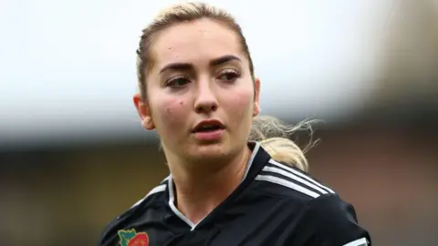 Getty Images Maddy Cusack playing for Sheffield United in 2021. Maddy has her long blonde hair tied back in a ponytail and wears a black football shirt with three white stripes on the shoulders. She's looking over her left shoulder with a serious expression. 