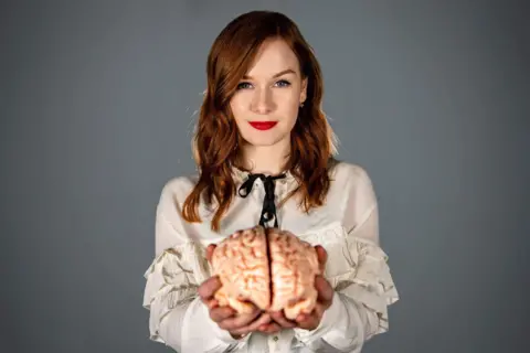 Daniel Harris Alice Gray holds up a model of a brain and smiles into the camera. She has long red hair and is wearing a ruffled white shirt.