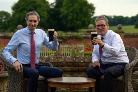 bbc two men in shirts and ties raising full pint glasses of Guinness in the direction of the camera. They are sitting in wicker chairs with roses and trees in the background, and are on either side of a round wooden table