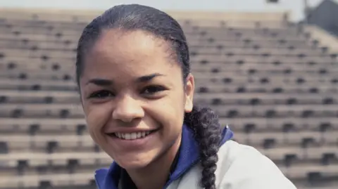 Getty Images A portrait of sprinter Anita Neil taken in June 1967 at the Crystal Palace National Sports Centre. Ms Neil, who is wearing a sports top and has her hair in a plait, is smiling.