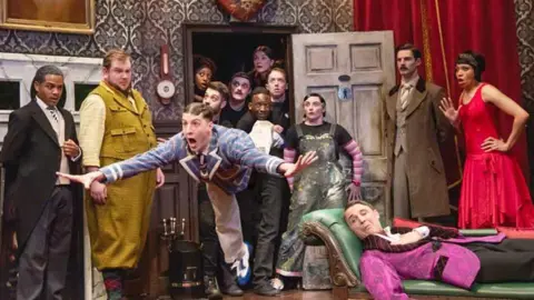 Matt Crockett An actor in The Play That Goes Wrong falls through the air as if he has tripped up while other cast and crew look on in shock. Another actor in a pink smoking jacket lies on a chaise longue looking worried