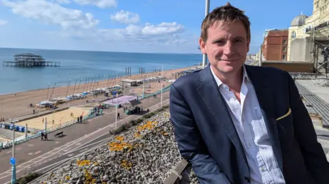 Jennifer McKiernan/BBC Mike Martin MP leans against a balcony overlooking Brighton Beach and Brighton Pier on a sunny day. He's wearing a navy suit, a pale open-necked shirt and his short, brown hair is spiked up a little in the middle 