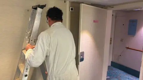 A maintenance worker holding a silver ladder whilst wearing white work overalls. There is a door open to the corridor.