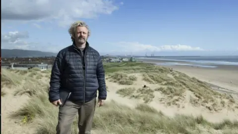 Jon Pountney The actor Michael Sheen stands on some sand dunes looking into camera wearing a navy padded jacket and holding a folder.