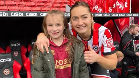 Contributor photo Neveah and Maddy on the day she was a Sheffield United mascot. Maddy, wearing her red and white striped kit, has her arm around Neveah's shoulder and gives a thumbs up to the camera. They are both smiling, Neveah missing some of her front teeth. 