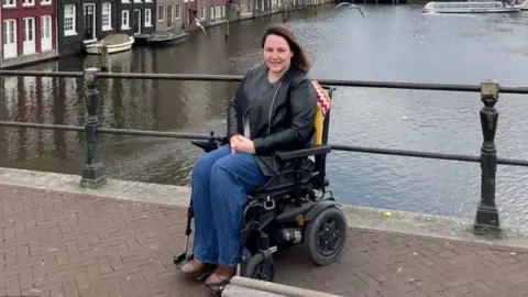 Sam Jennings Sam Jennings, who has long brown hair, wears brown boots, jeans and a black leather jacket. She is in a powerchair, with a canal behind her