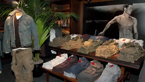 Getty Images An interior shot of the Abercrombie & Fitch store on 5th Avenue in New York City, taken at its opening event in 2005, showing piles of folded clothes, a mannequin dressed in a denim jacket and cargo trousers and with an advertising image of a shirtless model in the background