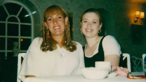 BBC/Joan Collins Gemma Collins and her mother, Joan. Both are sitting at a table and smiling at the camera. Joan is wearing a white shirt while Gemma is wearing a white T-shirt with a black top over it