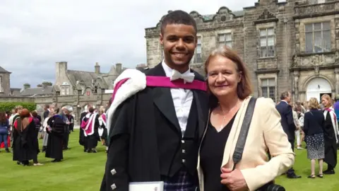 Christopher MacRae Christopher in kilt with black jacket, white shirt, white bow tie and graduation gown, at St Andrews University. He is standing next to his mum Sara, who is wearing a black top and cream jacket. Both are smiling.