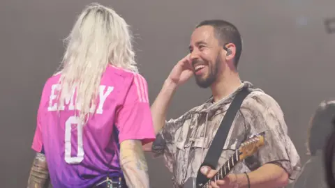 Getty Images Emily Armstrong and Mike Shinoda on stage in Hamburg as part of Linkin Park's From Zero world tour. Mike smiles at Emily, who's facing away from the camera, her long bleached hair loose down her back as she wears a pink sports shirt with her name and '0' on the back. Mike has cropped dark hair and a short beard and holds a guitar in one hand and his earpiece in the other.