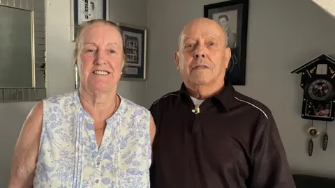 Tony Fisher/BBC A woman wearing a white and blue sleeveless blouse next to her husband who is wearing a black sports top.