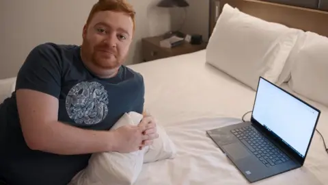 Bodhi Media/BBC A man with ginger hair and a beard lies on a bed in front of a laptop computer