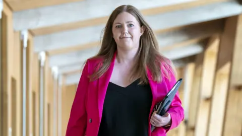 PA Media Gillian Mackay walks to the chamber in the Scottish Parliament, carrying a black folder. She is wearing a bright magenta jacket.