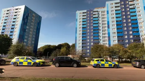 Emma Baugh/BBC Two police cars are parked in Marsh Farm outside the blue Leabank block of flats