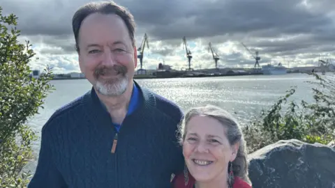 BBC Randy and Kit standing in front of Belfast's ship yard. Randy is wearing a navy zip-up fleece with a blue top and Kit is wearing a red cardigan with a black top underneath.